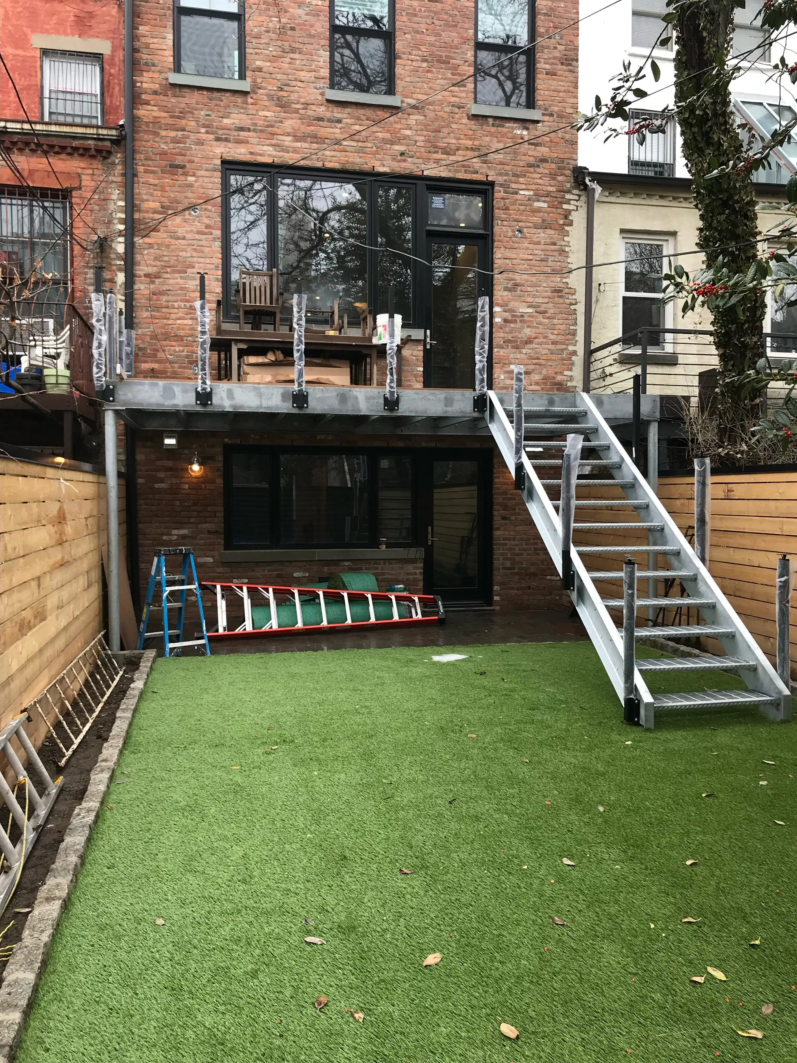Galvanized steel rear deck and open-riser stair attached to the back of a Brooklyn brownstone above a turf yard