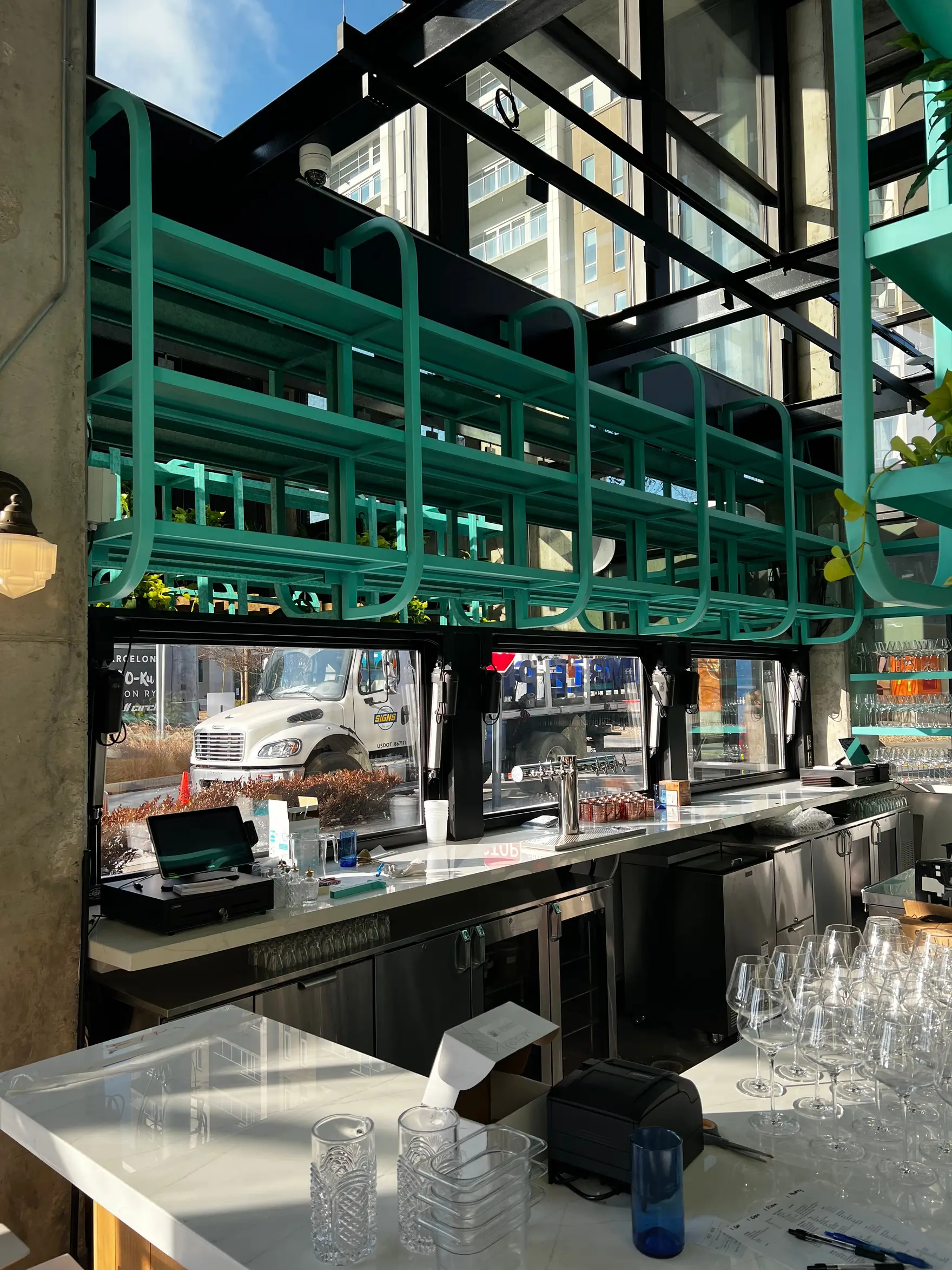 Teal powder-coated steel shelving frames suspended above a restaurant back bar beside a glazed façade