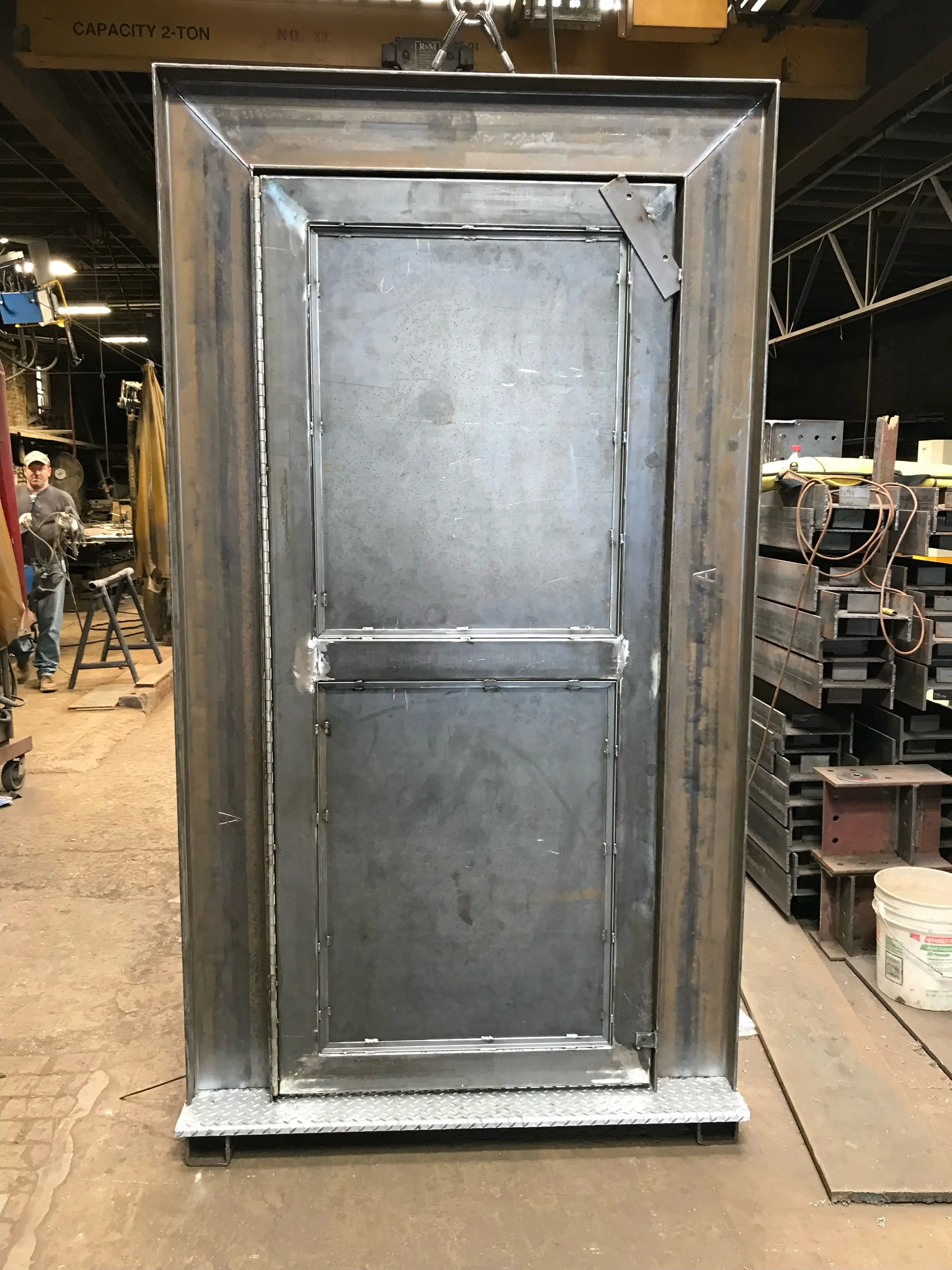 Welded steel security door and frame assembly hanging from a two-ton shop crane on the fabrication floor